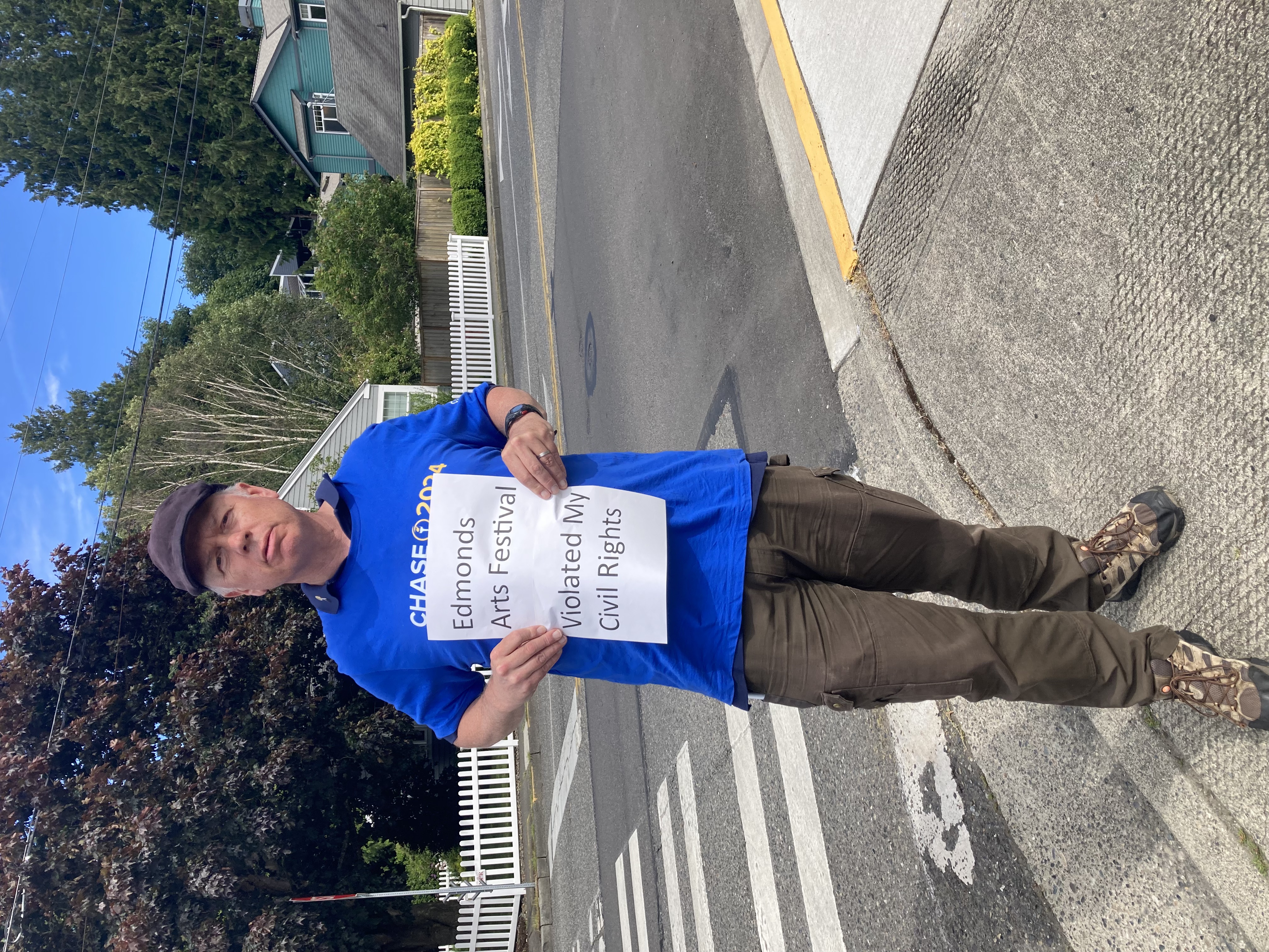 Bruce standing on a street corner holding a sign: "Edmonds Arts Festival Violated My Civil Rights"